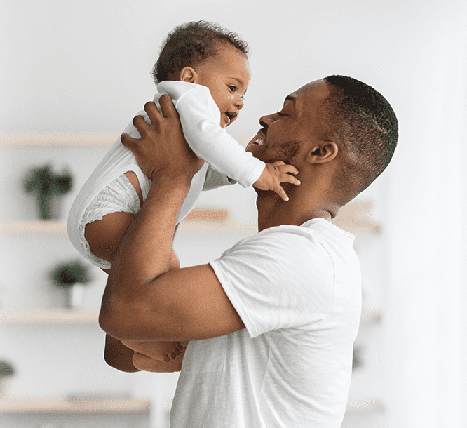 Um homem negro, com cabelo quase raspado, com uma blusa branca de manga curta, segurando uma criança negra, com cabelo curto, vestindo uma roupa branca e usando fralda. Os dois estão sorrindo.